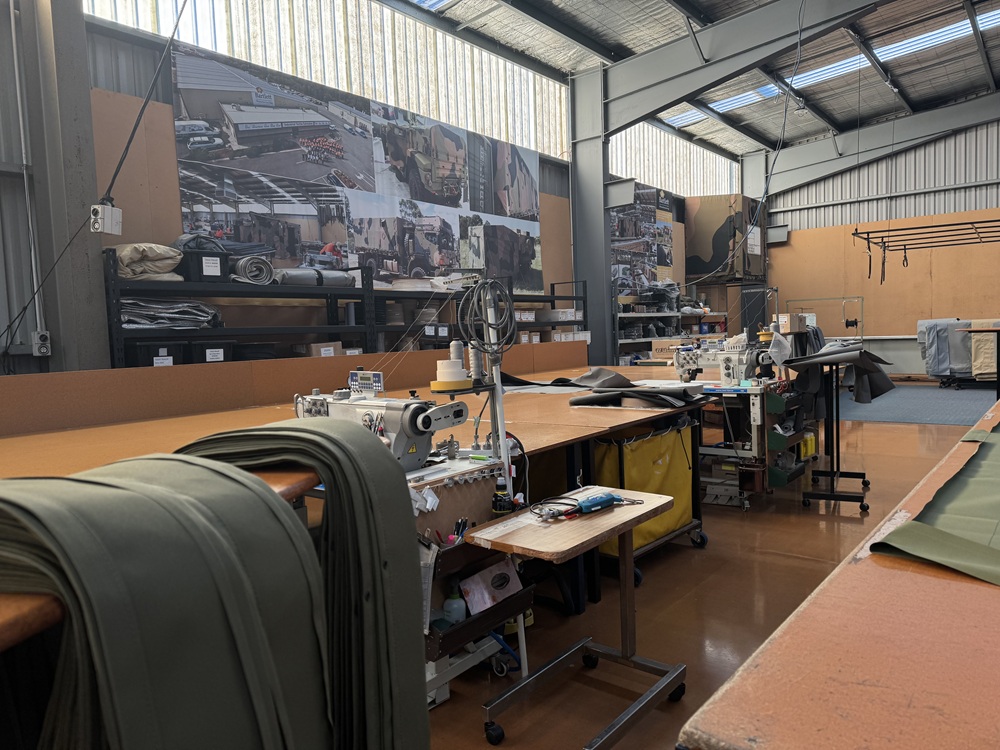 A white industrial sewing machine and brown table, with green fabrics layered to the left of the machinery. Similar stations are visible in the right side of the image, and pictures of the factory can be seen on the far wall. 