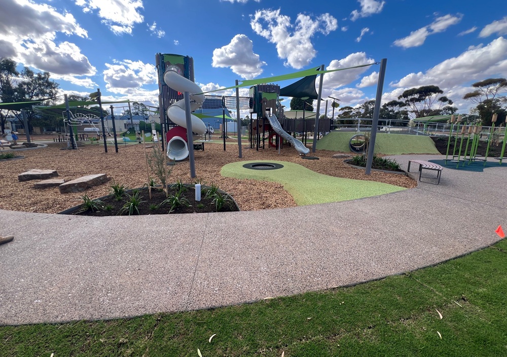 Various play equipment at the Robinvale precinct, including twisting slides, a tunnel and an in-ground trampoline, set under green shade sails and a tanbark play area. 