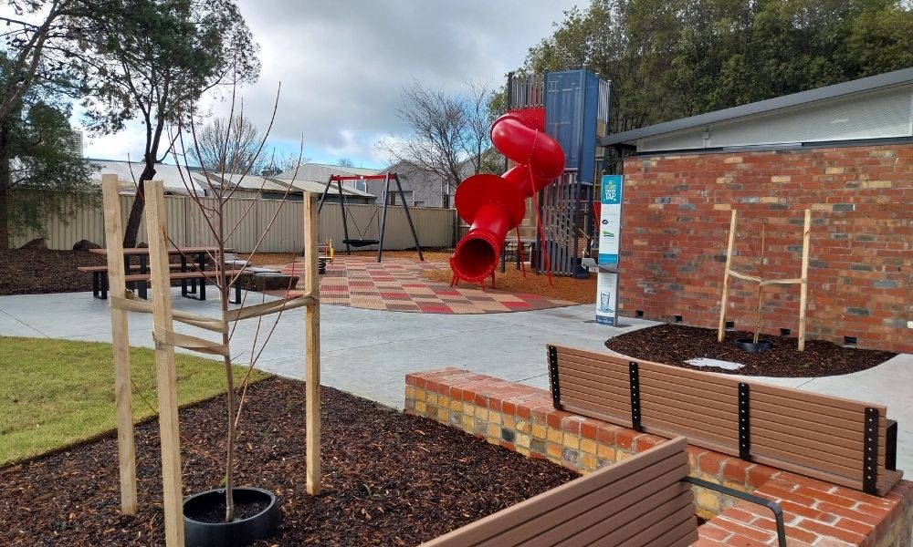 The playground and public space outside the Ballan Library and Community Hub. Public benches, a water fountain, tree plantings and a playground with a red slide and accessible swing are visible in the shot.